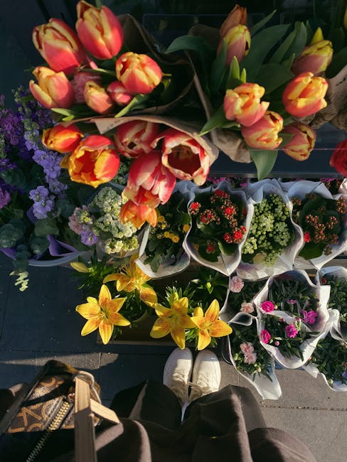 A vibrant display of freshly arranged flower bouquets and potted plants arranged on a outdoor surface, featuring a variety of tulips with yellow and orange petals, red and white tulips, yellow lilies,