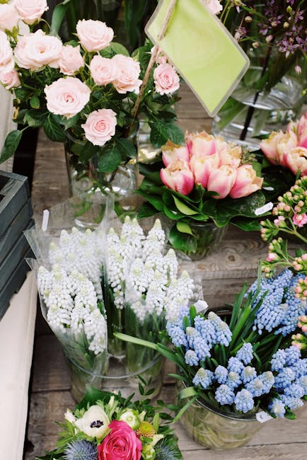 A variety of fresh floral arrangements displayed on a wooden surface, featuring a bouquet of pale pink roses with lush green foliage in a clear glass vase, adjacent to a bouquet of pink and cream tuli