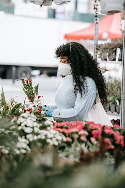 A woman with curly dark hair wearing a white face mask, light blue sweater, and white apron, arranging a bouquet of fresh flowers at an outdoor market stall. The bouquet includes red poppies, white li